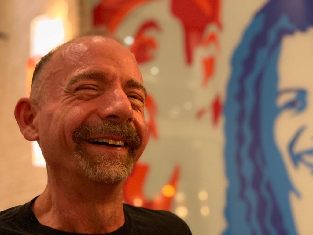 Timothy Ray Brown, conhecido como paciente de Berlim, sorri à frente de uma parede cheia de ilustrações. Ele foi a primeira pessoa no mundo a ser curada da infecção do HIV